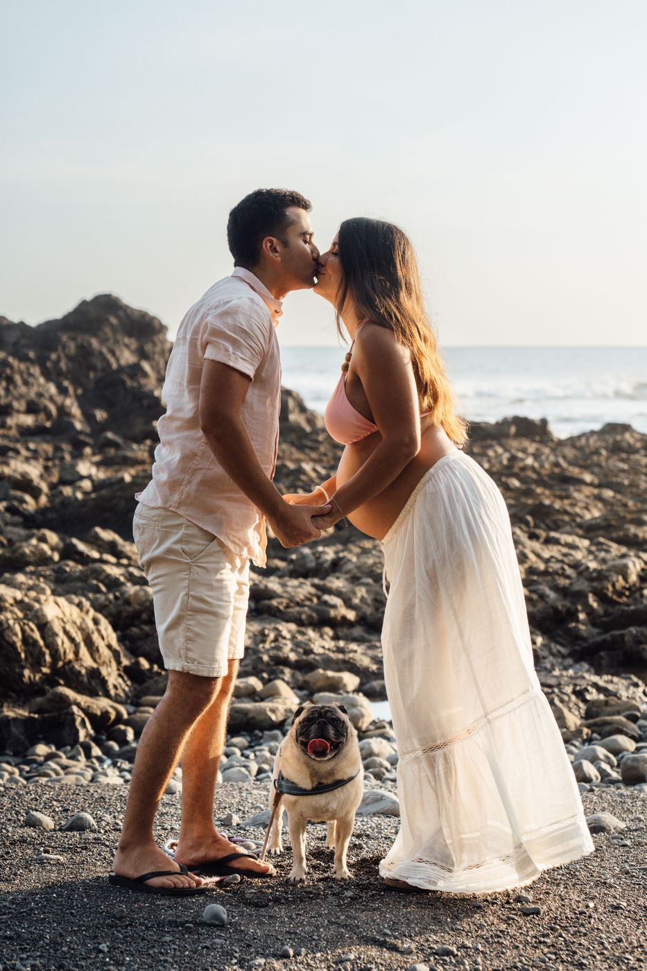 Pareja esperando bebé besándose en una playa de piedras, con su pug a los pies