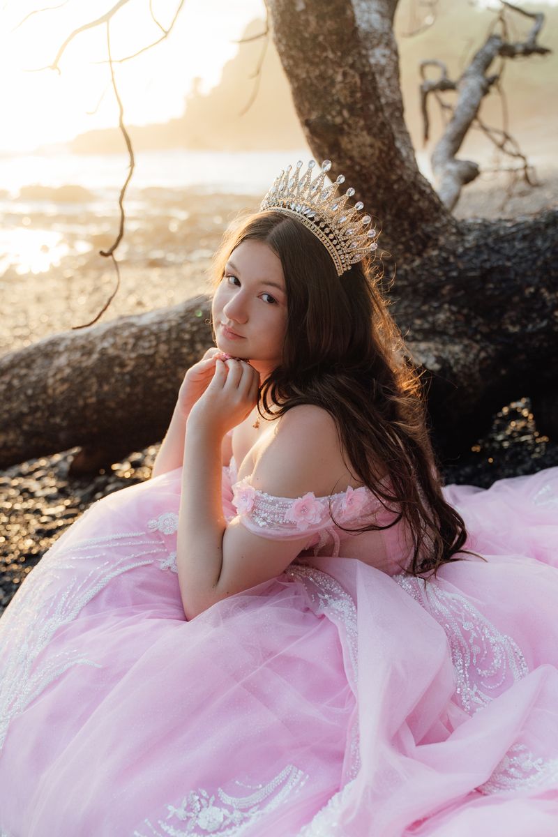 Quinceañera con vestido rosado y corona junto a un tronco en la playa